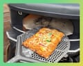 A rectangular pizza with melted cheese and toppings sits on a perforated metal tray in front of a pizza oven.