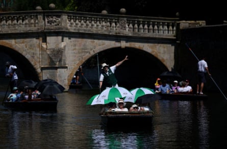 A man stands punting as people sit in a boat holding umbrellas. A stone bridge is in the background