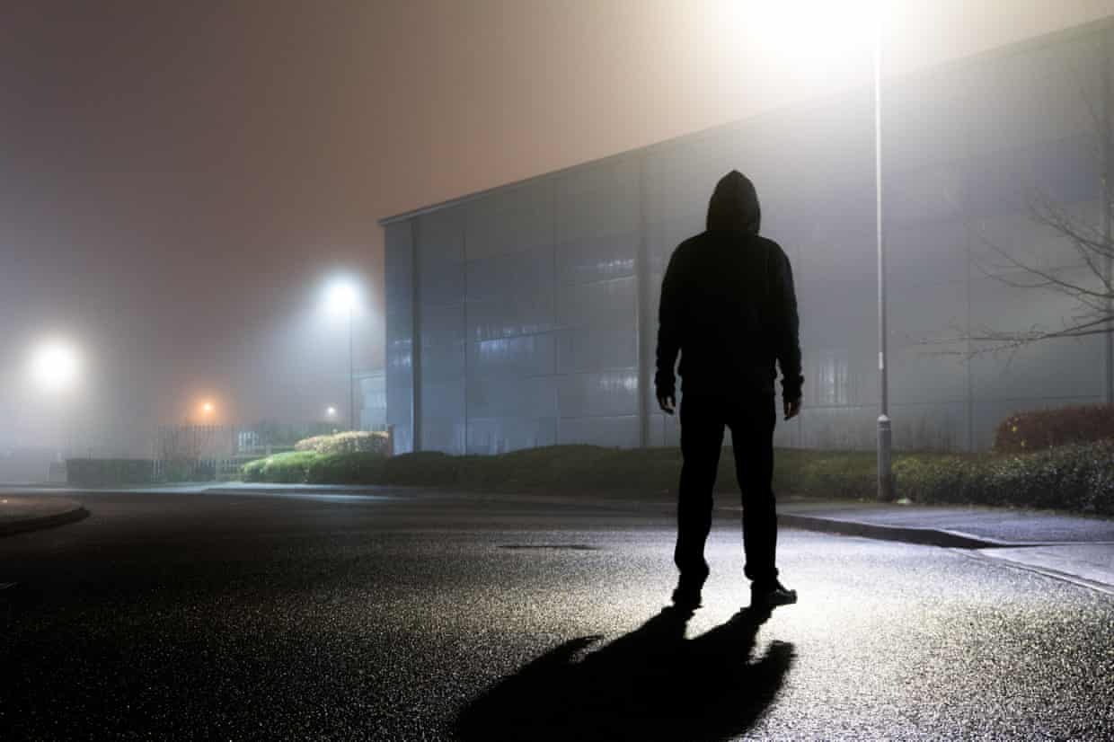 A sinister hooded figure, silhouetted by street lights on an empty industrial estate on a mysterious foggy night