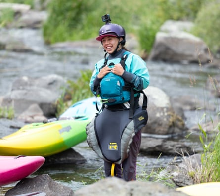Young woman in teal gear, helmet with camera on top, smiling alongside yellow and green kayak on a river.