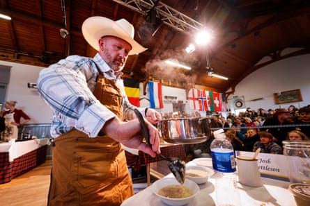 Josh Kettleson stirs a bowl on a table in front of the crowd in the hall. He wears a brown apron, a checked shirt and a cowboy hat. National flags representing the competitors hang on the wall at the back of the hall.