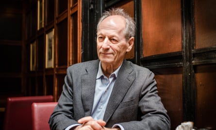 Michael Marmot pictured in a dark wood-panelled interior on a red leather seat, with his hands folded on a table. He wears a grey suit and open-necked pale blue shirt; he has thin, receding grey hair and has a small smile but looks serious.