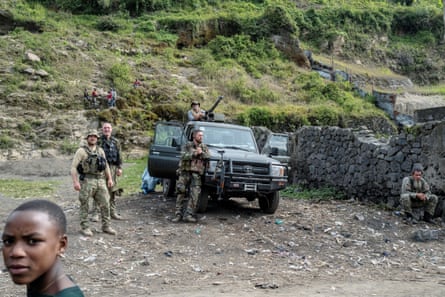 A group of white men in combat gear and holding rifles stand around a military 4x4 vehicle