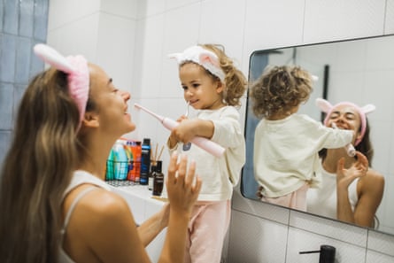 A toddler stands up at the sink and brushes her mumâÂÂs teeth with an electric toothbrush. Both are wearing fluffy bunny ears