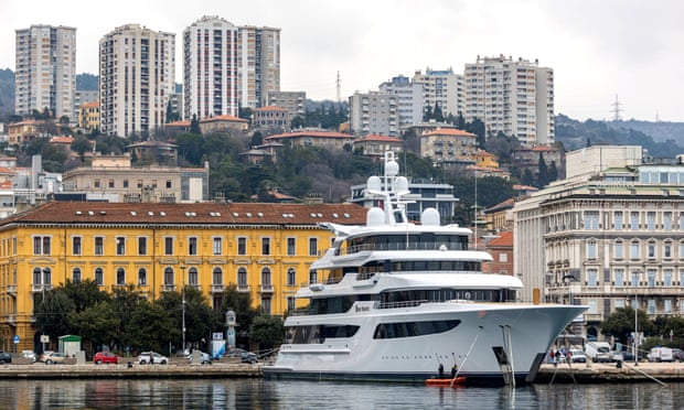 A large white yacht sits at a dock with a cityscape in the background.