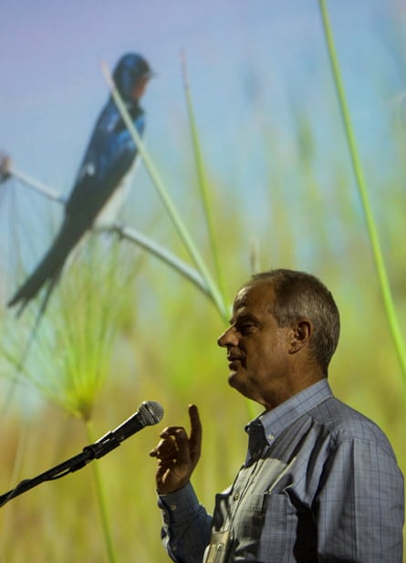 An older white man in a collared shirt speaks into a microphone and gestures at a projected image behind him of a blue-colored, red-throated bird perching on grasses