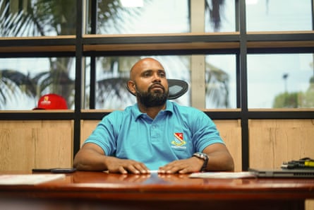 Miguel Angel Castell Cano sits at a desk in his office in front of a window, with palm trees seen outside. He wears a blue polo shirt and has a neat beard and shaven head.