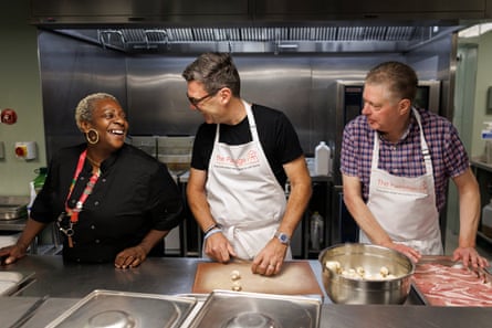 Burnham wearing apron, laughs with woman, as he and man on his other side prepare food