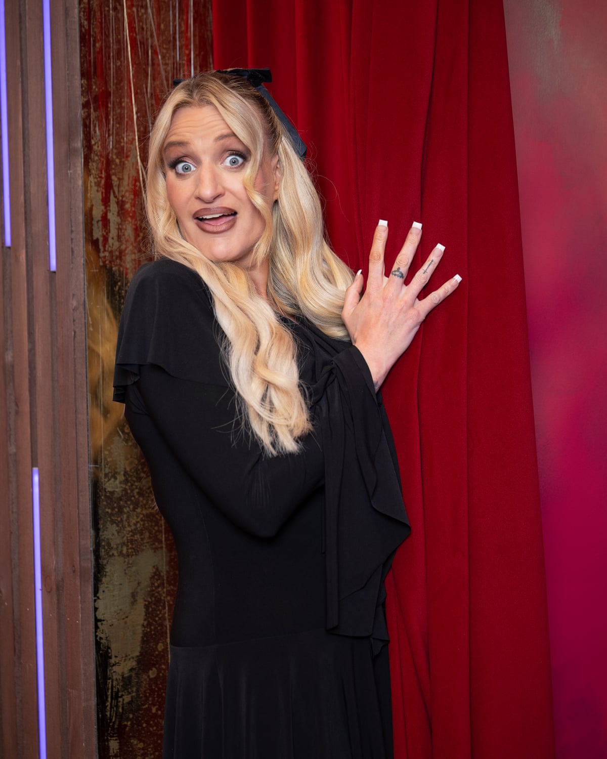 Daisy May Cooper peeks out from behind a red curtain with a surprised expression