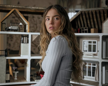 A woman standing beside a doll’s house, looking over her shoulder