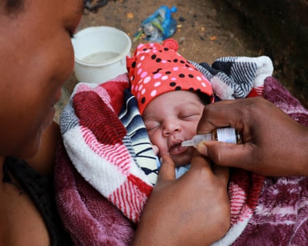 A baby is being held as a hand holding a medicine dropper reaches towards its open mouth.