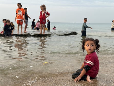 A small girl kneels on the sand at the water's edge as older children stand in the water or on a rock