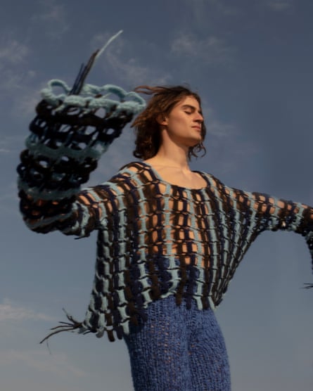 A model in a blue and black crocheted jumper with wide sleeves, against a blue-grey sky.