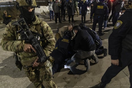 A federal agent in a camouflage uniform and helmet stands holding an assault rifle at the ready, his face covered up. Behind him, a group of law enforcement officers hold a protester on the ground.