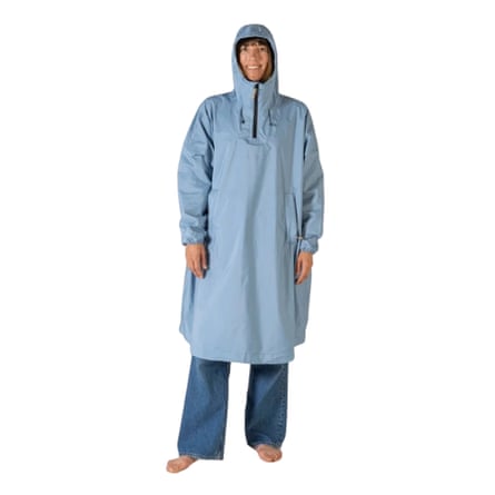 A smiling woman wears a long light blue hooded rain poncho over jeans, standing barefoot