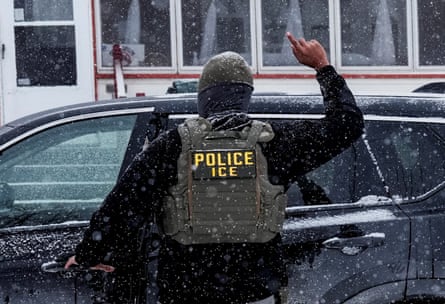 Masked agent raises a finger with his other hand on a vehicle door handle in snowy conditions