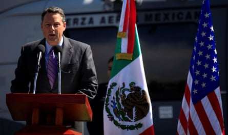John Feeley addresses an audience from a stage behind microphones. He stands in front of Mexican and US flags.