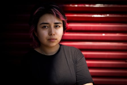A girls with a pink streak in her dark hair stands in front of a red shutter.