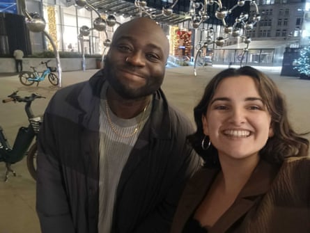 A man and a woman smiling while they take a selfie standing outside with a large silver metal sculpture behind them