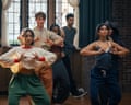 Maya and Sumi lead a group dance move, hands crossed over chests, others following behind