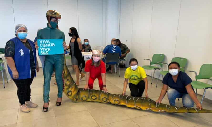 Klinger Duarte Rodrigues stands next to a health worker at a vaccine clinic wearing a costume to look like an Amazonian water boa and holding a sign celebrating science and the Brazilian health care system. Three people are holding his "tail".