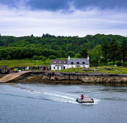 A boat travelling on the water, leaving the shore. Two small buildings are in the background amongst hills of greenery.