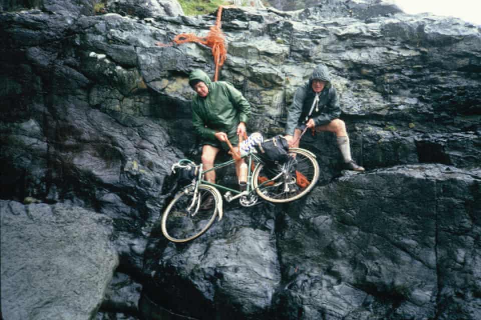 Two cyclists lower a bike down a rocky face.