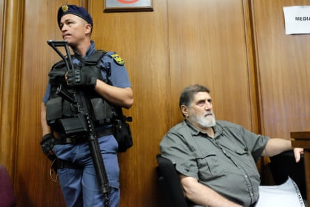 Eugene de Kock sitting in front of the wood panelling of a courtroom with an armed police guard standing next to him