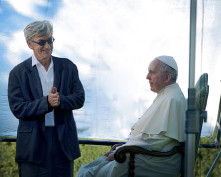 Wim Wenders, in a suit and shirt and sunglasses, pushes both hands together as he looks at the pope seated in front of a canvas outside