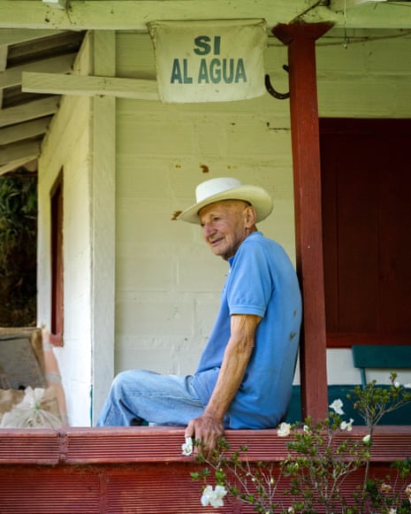 An older man in a hat sitting on wooden porch of a farm house.