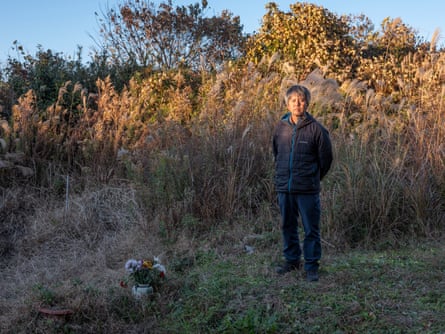 A Japanese man stands in a clearing among bushes, a vase of flowers on the ground beside him