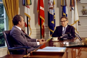 Nixon sitting at a desk in the Oval Office with Kissinger sitting in front of flags