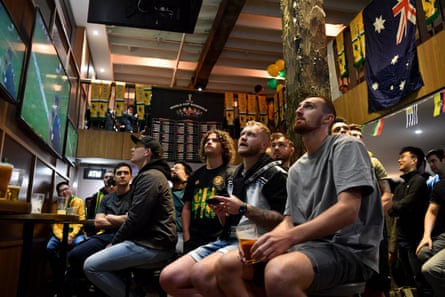 Supporters sit on stools in a pub watching a football game on tv screen while Socceroos and Australian paraphernalia hang on the walls