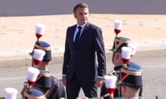 Emmanuel Macron at a Bastille Day parade in Paris, 14 July 2024: he is seen in the centre of the picture in a street with lines of soldiers in ceremonial uniform and plumed hats facing him. He wears a dark blue suit with shirt and tie and is positioned so he appears to be looking down the ranks from a height.