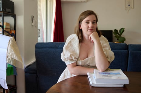 Maddy Hoffman is a Perth PhD student who has had to rely on her partner's income to get her over the line. She is wearing a neutral-coloured dress and sitting at a dark wooden table with two books in front of her
