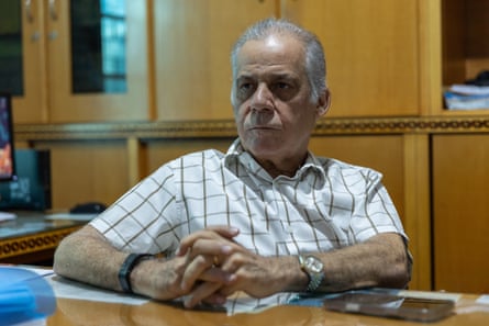 Portrait of an older man in a white, checked shirt sitting at a desk