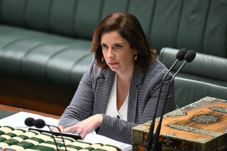 Anika Wells, wearing a black and white blazer and her shoulder length hair parted on the side sits behind two microphones and in front of a green leather bench