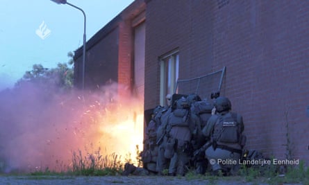 Photo of police huddle in a group against the wall of a building as a large explosion goes off by the door