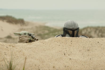 Grogu with a mini telescope and the Mandalorian tucked behind a sand dune