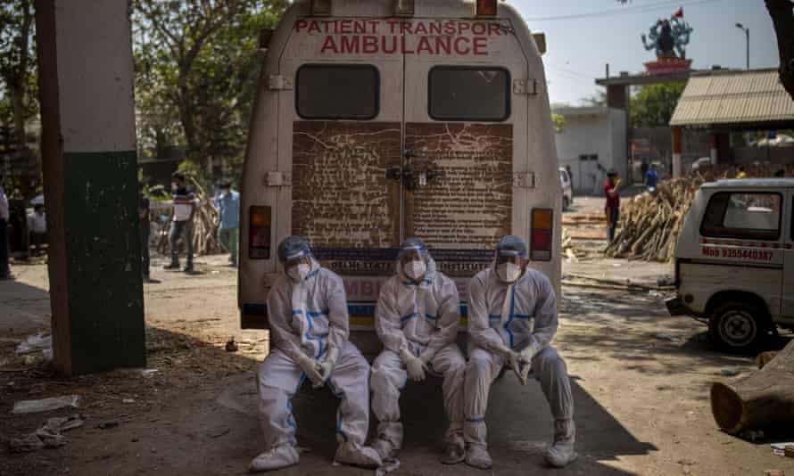 Three workers in white PPE sit on the back step of an ambulance at a crematorium in New Delhi