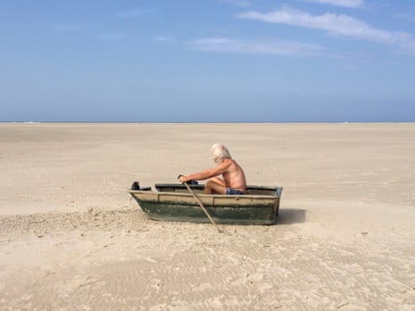 A man appearing to row a wooden boat sitting on a large expanse of sand