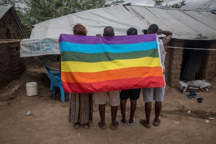 Four Africans in front of a small shelter with a tarpaulin roof holding a rainbow LGBTQ+ flag over their shoulders