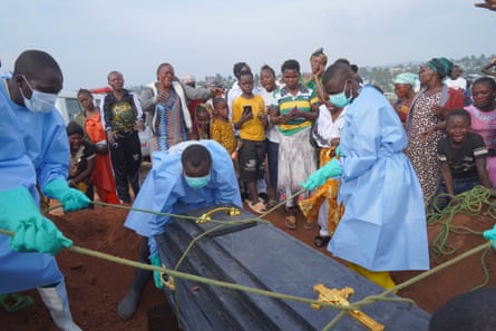 Three men in protective gear lower a coffin into the ground as mourners stand around the grave