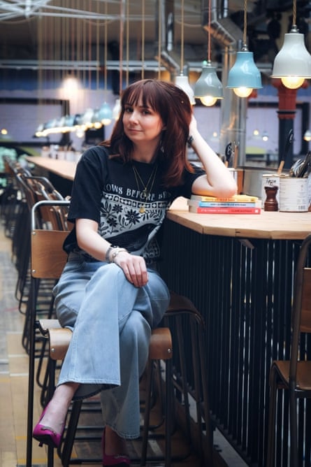 Claire Eastham sitting on a bar chair with head resting back on hand. wearing blue jeans and black logo T-shirt