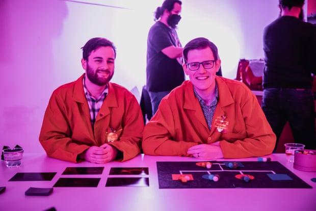 Two scientists travel with LED badges at the Beaker Street Festival. Stem cell explorer Alastair on the left and gene hunter Nicholas on the right. They sit in front of their desks with diagrams and pictures.the room is bathed in pink light