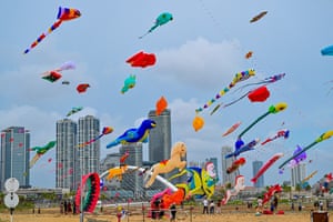 People on a beach fly multicoloured kites, some in the shape of animals, with high-rise buildings in the background