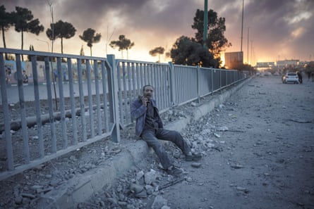 A confused-looking man sits by railings as smoke billows in the background