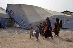 Three women in chadors and their children walk past an abandoned tent that has been blown on top of another one by the wind