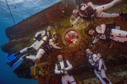 Six divers around the porthole of a submersible in ocean. Two people are looking out the window from inside