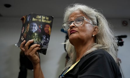 Grim-faced woman holds up book, the cover page and title visible